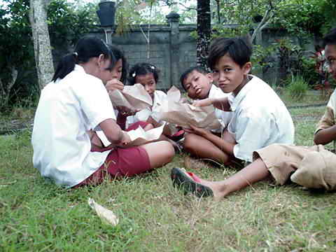 Mehrere Kinder sitzen auf der Wiese und essen eigepackten Reis mit Beilagen.